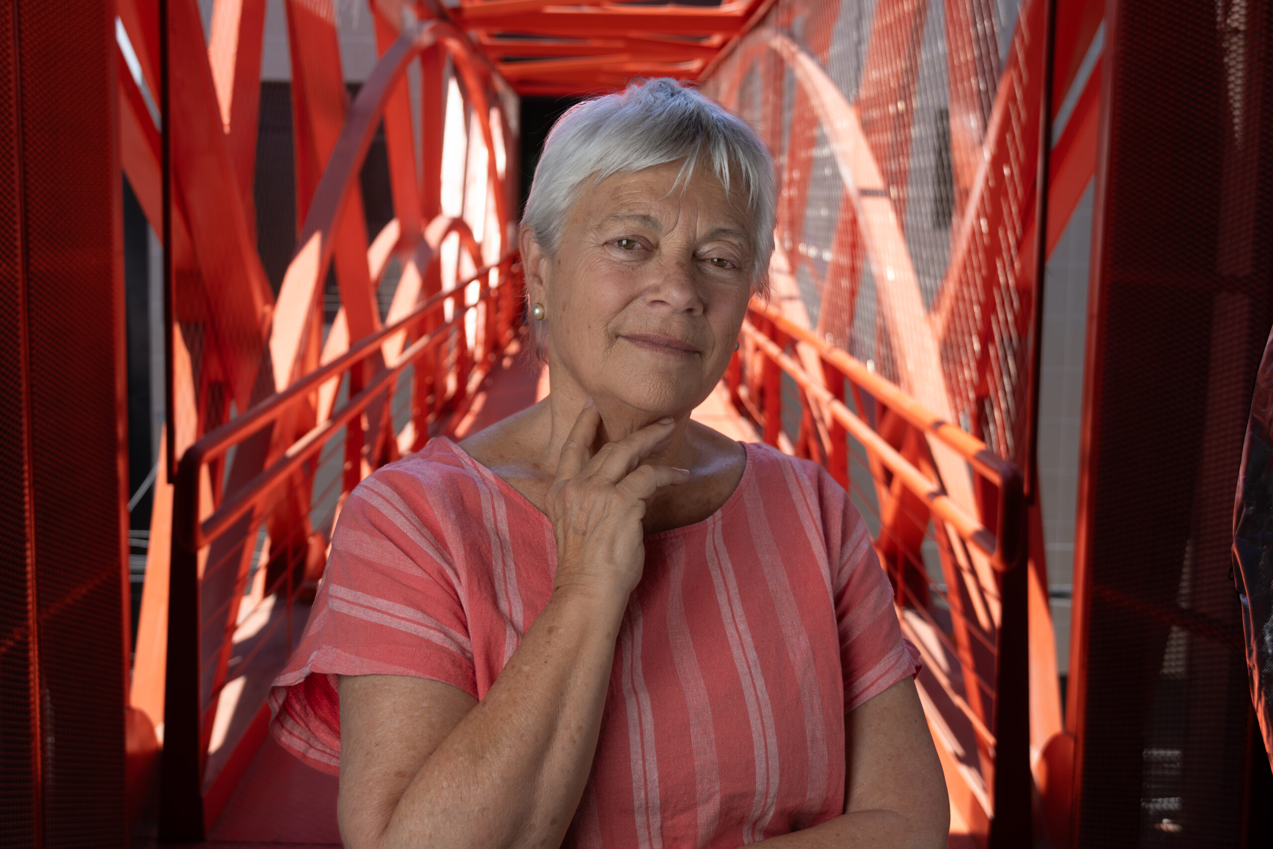 "Andrea Zondek, de cabello canoso corto, posa con expresión serena sobre una pasarela roja de estructura metálica."
